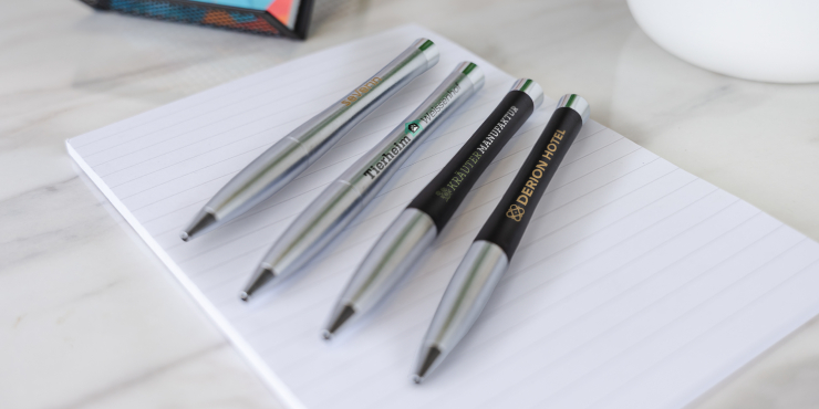 Vier Urban Classic Kugelschreiber von Parker mit eigenem Design liegen auf einem weißen A4-Papier neben einem Haftnotizblock und einem weißen Blumentopf