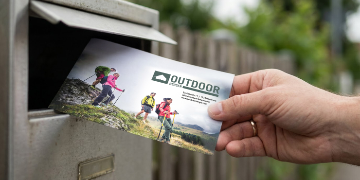 Eine Hand wirft gerade ein personalisiertes Briefcouvert in einen silbernen Postkasten 