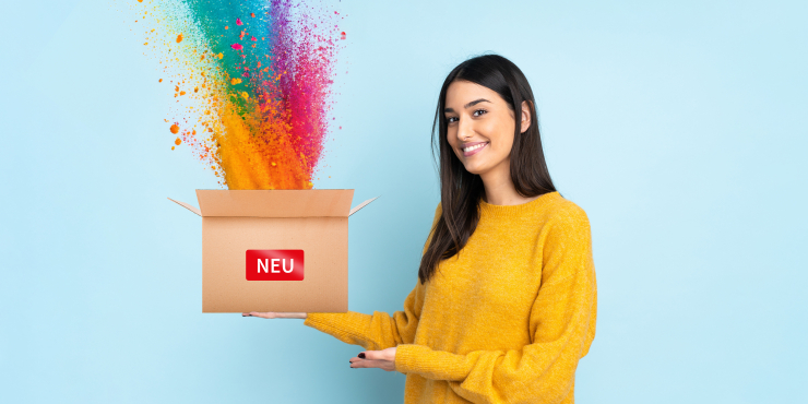 Frau im gelben Pullover steht mit Box, wo Neu oben steht und Farben herausspritzen, vor einem hellblauen Hintergrund 