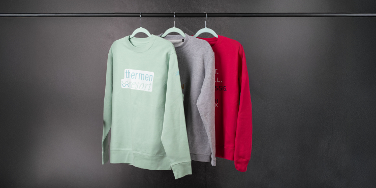 Mintgrüner, grauer und roter Sweater gestaltet mit eigenem Logo hängen auf einer Stange