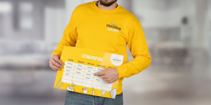 Lehrer zeigt auf Stundenplan und trägt gelben B&amp;C™-Sweater mit Aufdruck auf der linken Brustseite