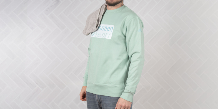 Männliche Person steht vor einer Fliesenwand und hat einen mintgrünen Stanley/Stella-Hoodie an mit personalisiertem Aufdruck auf der Brust