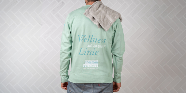 Männliche Person steht vor einer Fliesenwand und hat einen mintgrünen Stanley/Stella-Hoodie an mit Aufdruck am Rücken