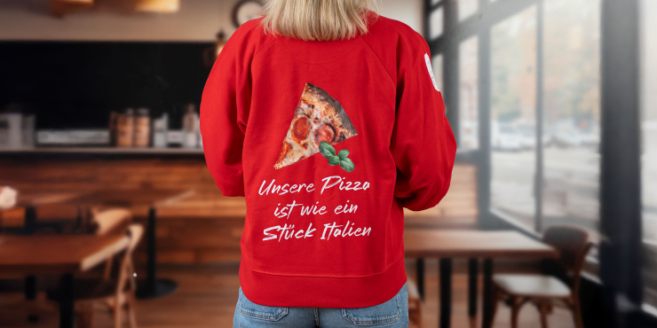 Weibliche Person steht in der Pizzeria und trägt einen roten Stanley/Stella-Sweater mit kreativem Aufdruck auf dem Rücken