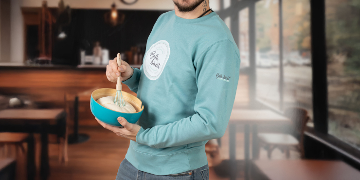 Männliche Person steht in der Backstube mit Schneebesen und Schüssel und trägt einen hellblauen Stanley/Stella-Sweater mit personalisiertem Aufdruck auf der Brust und auf dem Ärmel