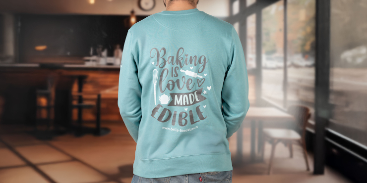 Männliche Person steht in der Backstube und trägt einen hellblauen Stanley/Stella-Sweater mit personalisiertem Aufdruck auf dem Rücken