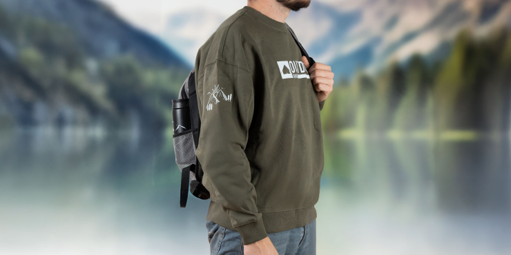 Männliche Person geht auf Wanderung, trägt einen Rucksack und einen olivfarbenen Stanley/Stella-Sweater mit Aufdruck an der Brust