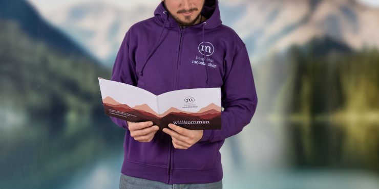 Männliche Person liest Broschüre in der Natur und trägt lilafarbenen Zip-Hoodie mit Aufdruck auf der linken Brustseite