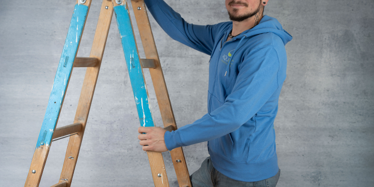 Heimwerker klettert die Leiter hinauf und trägt einen blauen Stanley/Stella-Zip-Hoodie, der auf der linken Brustseite ein Logo aufgedruckt hat