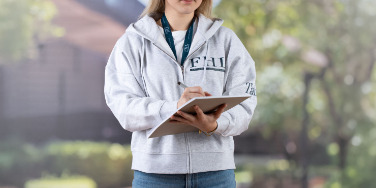 Studentin in grauem Zip-Hoodie bedruckt mit Logo steht am Campus und macht Notizen auf einem Schreibbrett