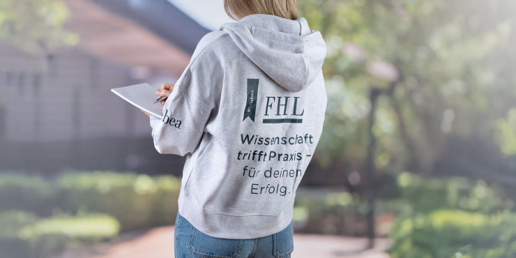  Studentin, die einen grauen Stanley/Stella-Zip-Hoodie mit Druck auf der Rückseite trägt, steht am Campus und macht Notizen