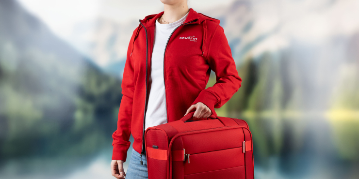 Weibliche Person mit rotem Koffer geht in der Natur und hat einen roten Zip-Hoodie mit Logo an