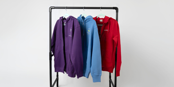 Dunkellilafarbener, hellblauer und roter Zip-Hoodie mit verschiedenen Logos hängen auf einer schwarzen Kleiderstange