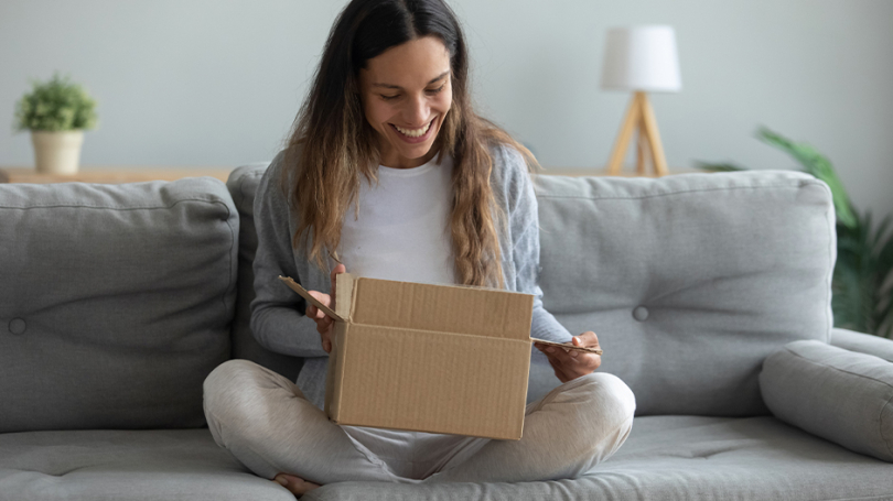 Eine Dame sitzt auf einem Sofa im Schneidersitz und öffnet einen personalisierten Versandkarton.
