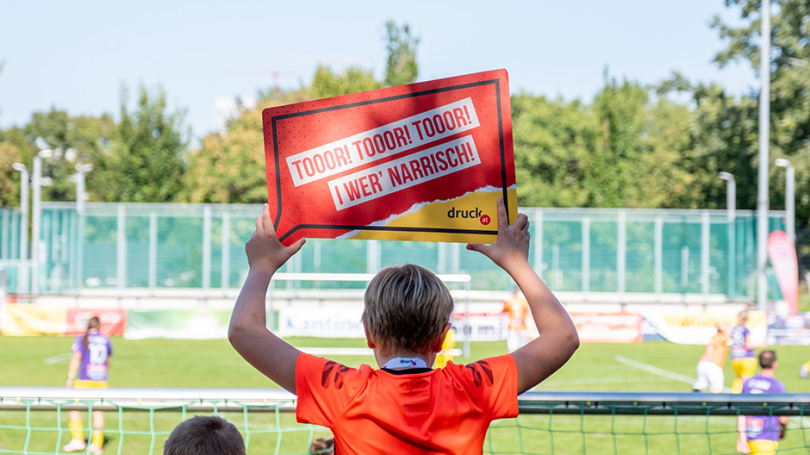 Ein Junge hält ein Anfeuerungsschild von druck.at hoch und feuert beim BFC an